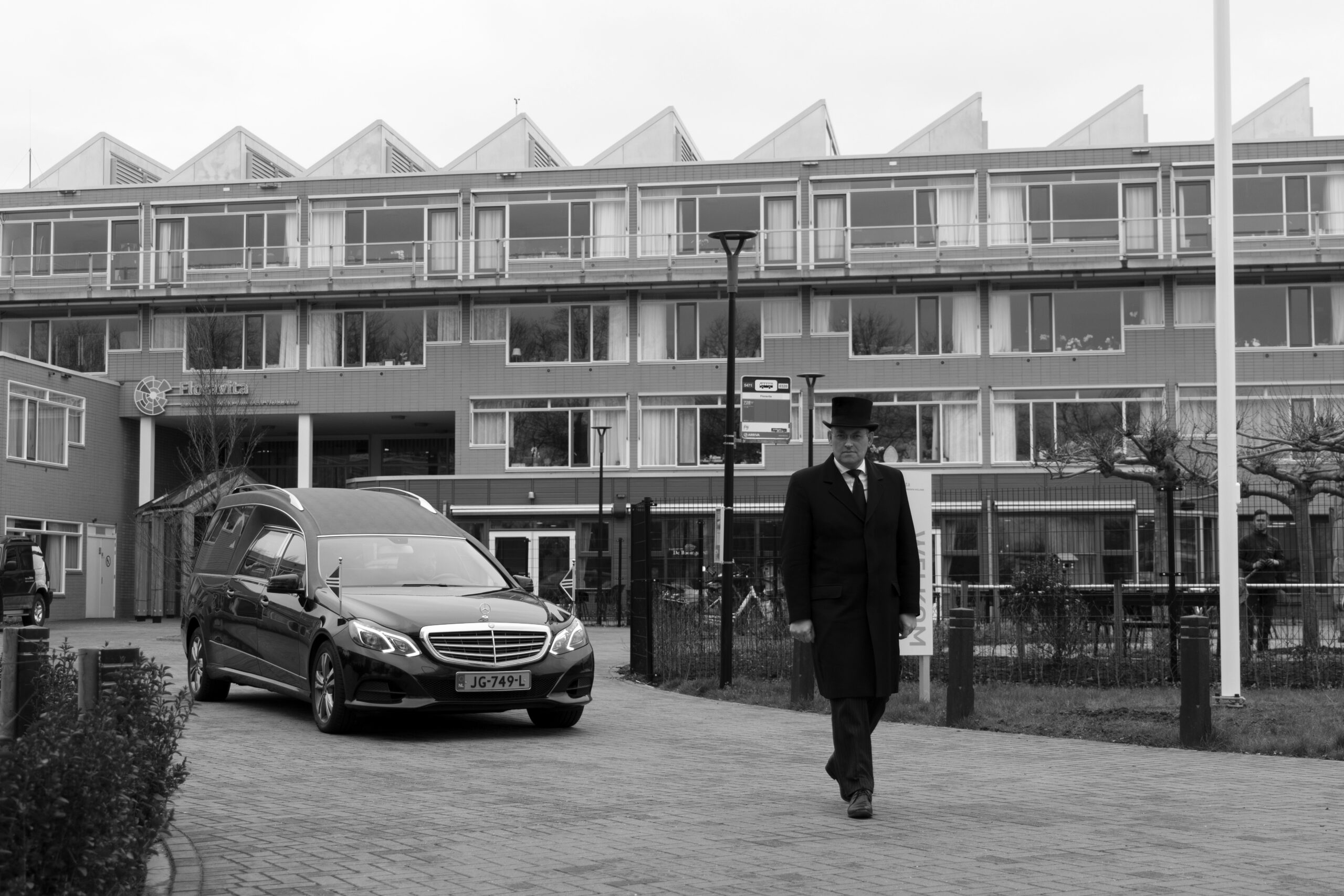 Uitvaartbegeleider loopt voor rouwauto die zorgcomplex verlaat